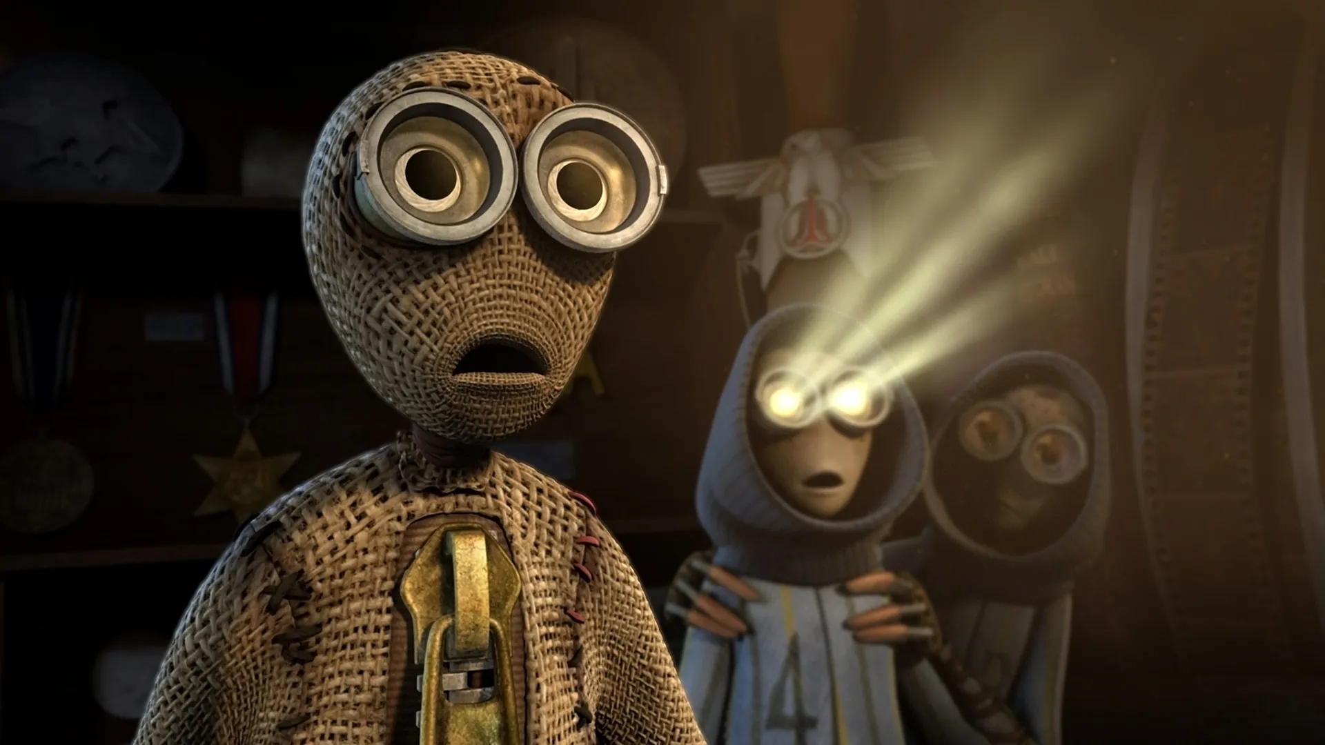 A close-up of a burlap-textured, humanlike robot with large round goggles and a stitched mouth, staring forward in a dim room. Behind it, another small robot with a glowing yellow headlamp sits between shelves and tools, casting a soft beam of light while a third robot with a circular face is partially visible in the shadows.